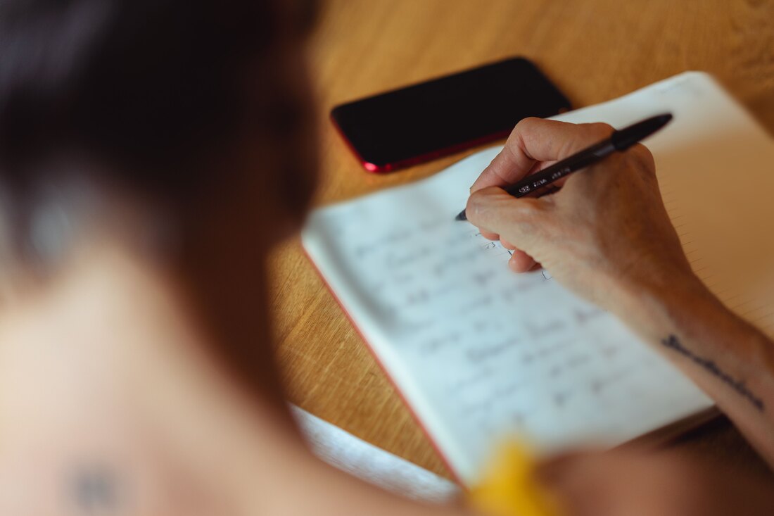 A hand writing in a notebook at a wooden table, a phone beside it