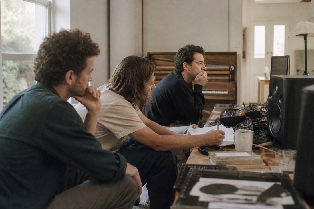 Three musicians listening intently to a track on studio monitors, one taking notes