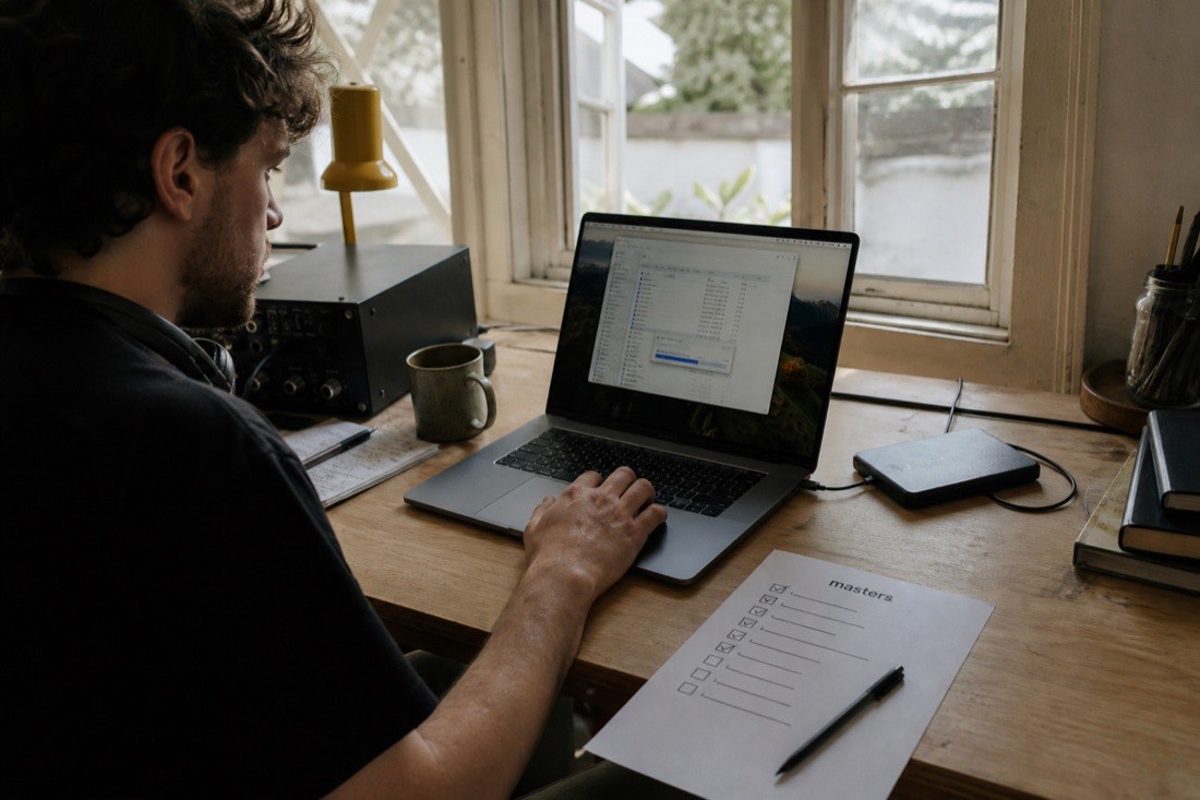 A musician at a desk in afternoon light, moving files from a laptop to an external drive, with a printed checklist titled masters half ticked off