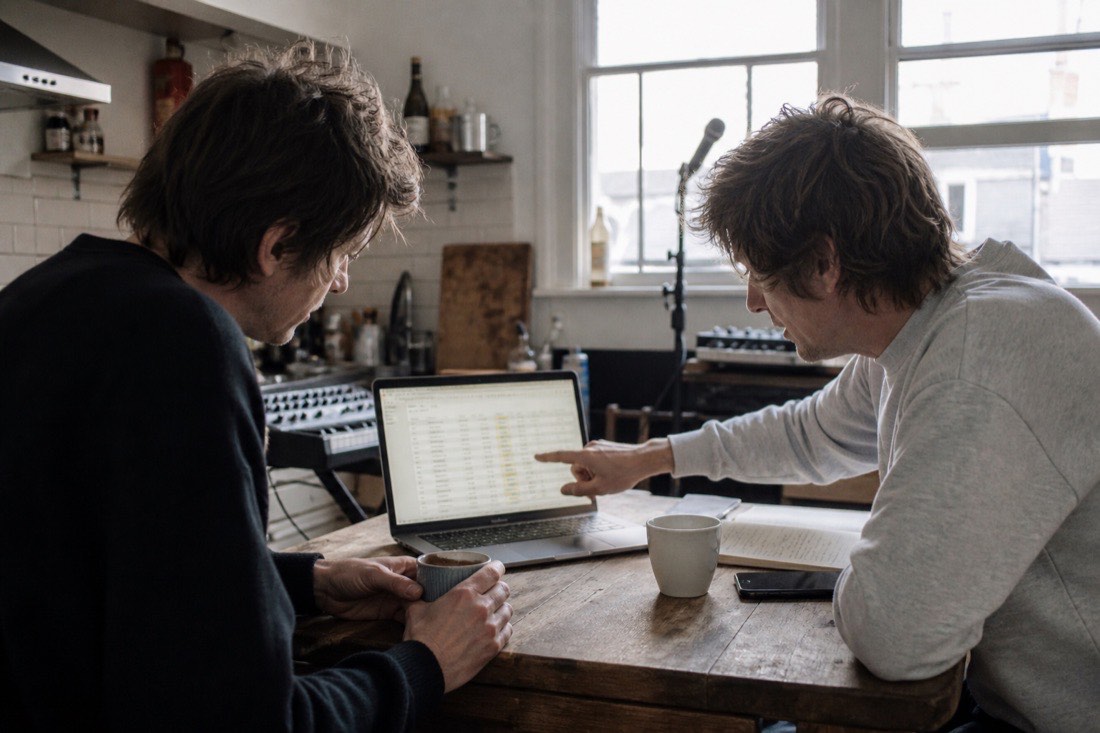 Two bandmates in a daylit studio kitchen, one holding a laptop showing a comparison table, the other leaning in and pointing at a line, coffee cups on the counter