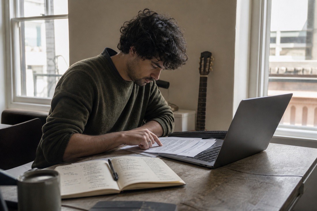 A musician alone at a desk in daylight, reading fine print on a laptop, one finger tracing a printed terms page beside a notebook with two columns of handwritten pros and cons