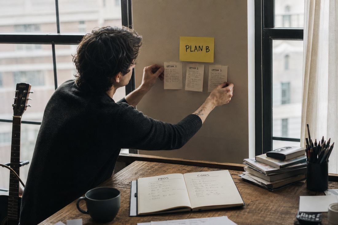 A musician by a big window pinning three option cards under a sticky note, notebook open with pros and cons columns, focused not defeated