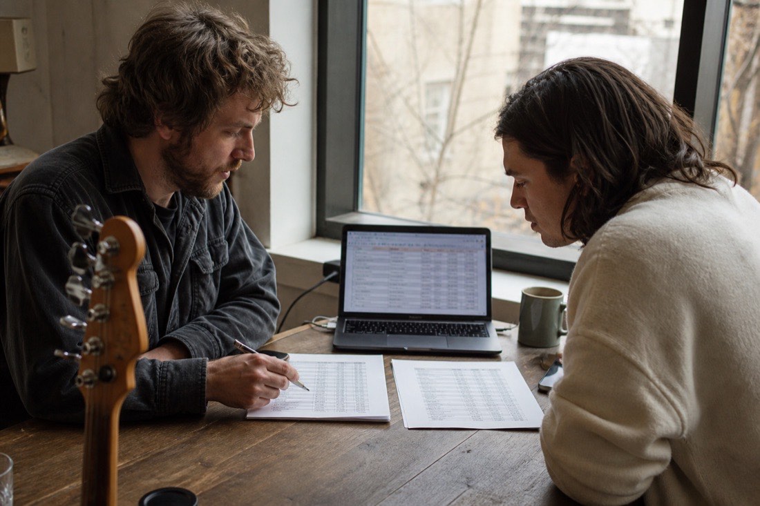 Two musicians at a table by a window comparing printed pricing sheets side by side, one annotating with a pen, a laptop open beside them