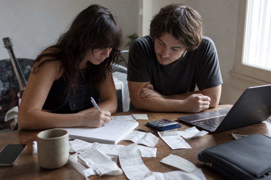 Two musicians at a table in daylight itemising release costs on a paper pad, calculator and receipts beside an open laptop