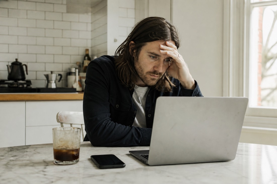 A musician at a kitchen table in daylight staring at a laptop, one hand on their forehead, phone face-down beside a cold coffee — tense but composed