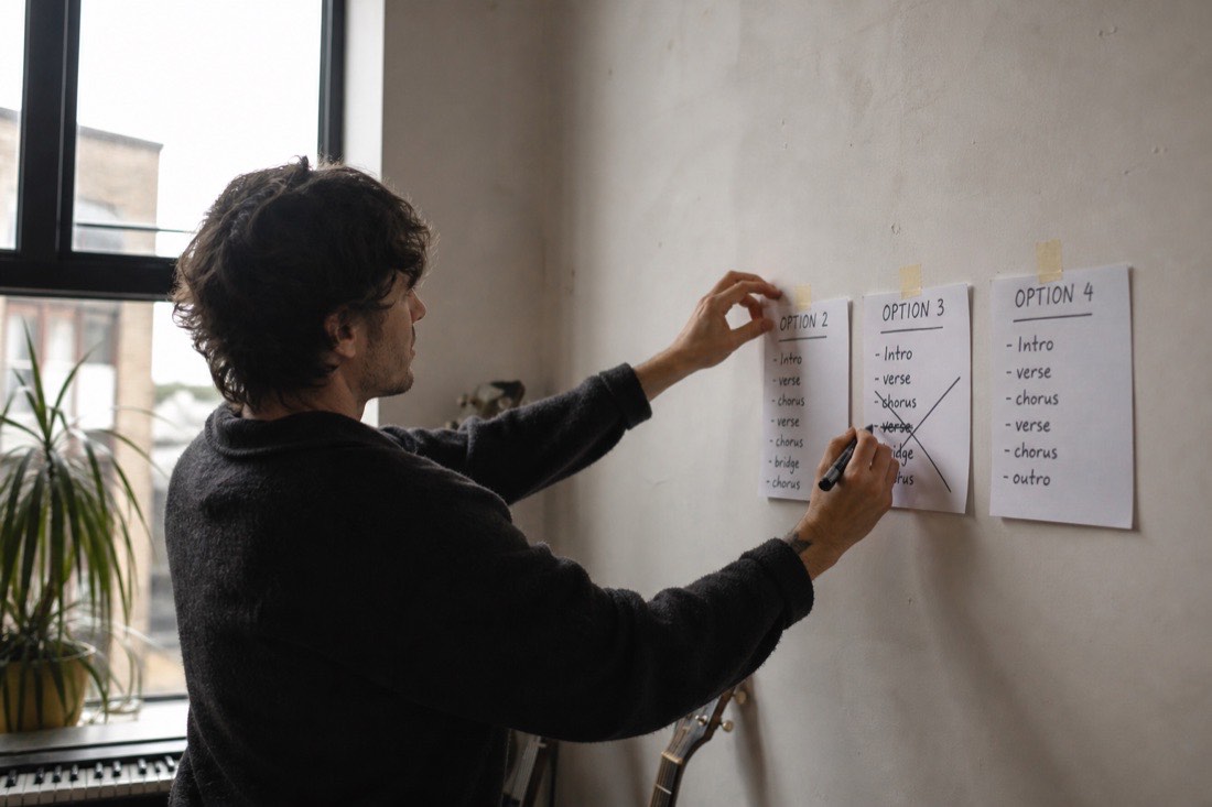 A musician taping four handwritten option cards to a wall in daylight, marker in hand, one card crossed out