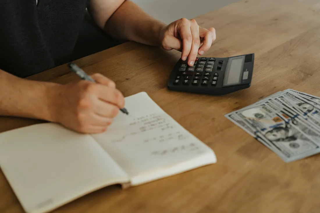 Hands at a wooden table with a calculator, an open notebook and a few US dollar bills