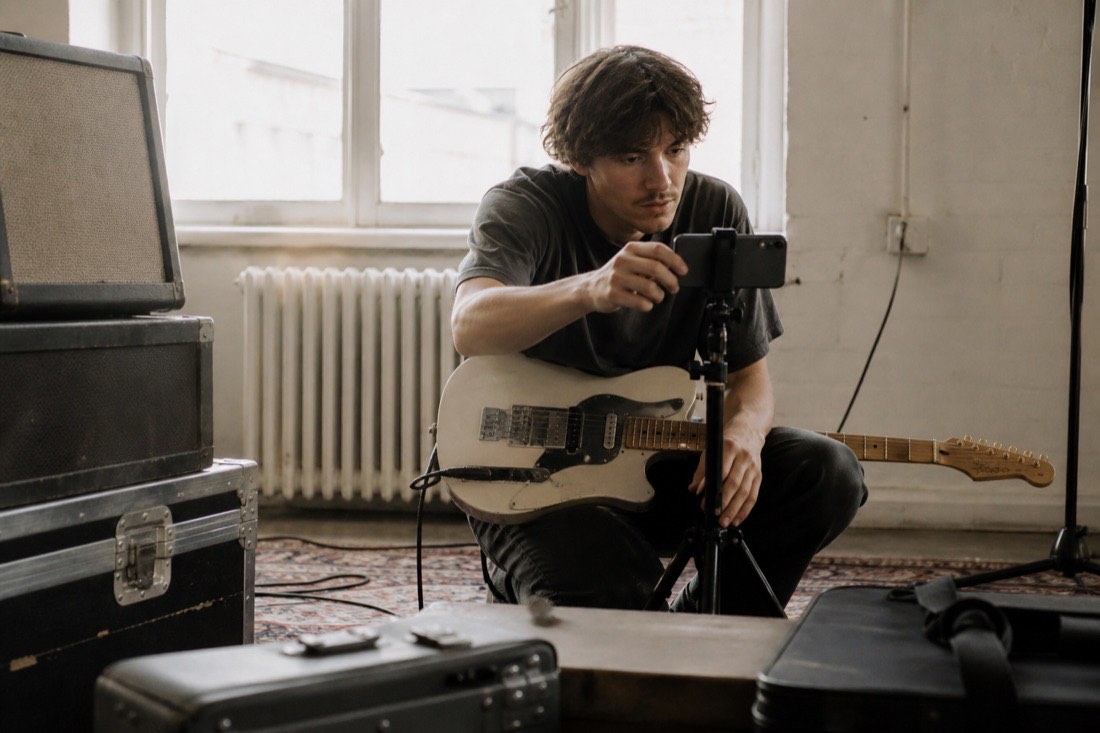 A musician in a daylit rehearsal room propping a phone on a tripod to film themselves playing, cables and amp cases around