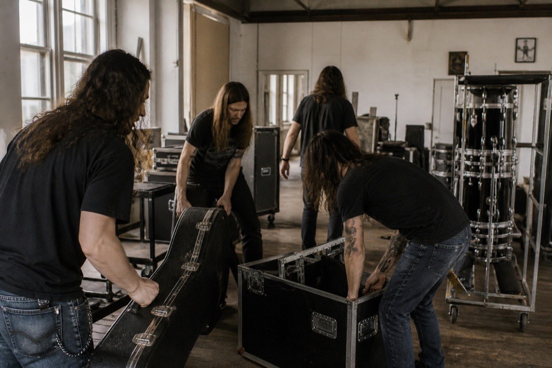 A metal band loading guitar cases and a drum rack into a daylit rehearsal space