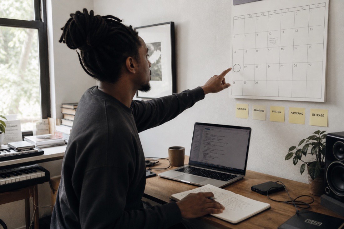 A musician at a daylit desk checks a wall calendar with a circled release date three weeks out, laptop open, a row of sticky notes marking the stages