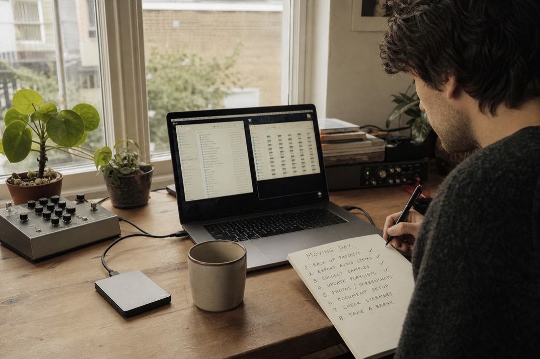 A musician at a daylit desk with two browser windows open side by side on a laptop, an external drive plugged in, and a handwritten checklist of numbered steps half ticked