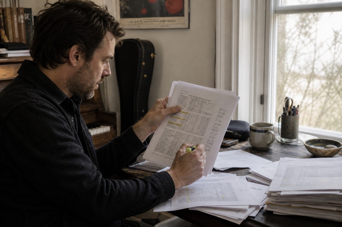 A musician at a desk comparing a stack of printed yearly invoices, highlighter in hand, sums written in a margin, window light