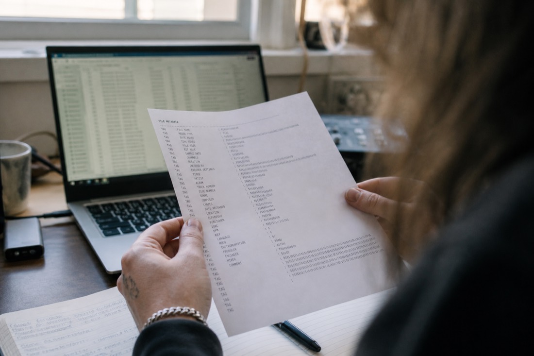 Close over-shoulder view of a musician's hands holding a printed metadata sheet with rows of codes, a laptop with a spreadsheet blurred behind, morning light