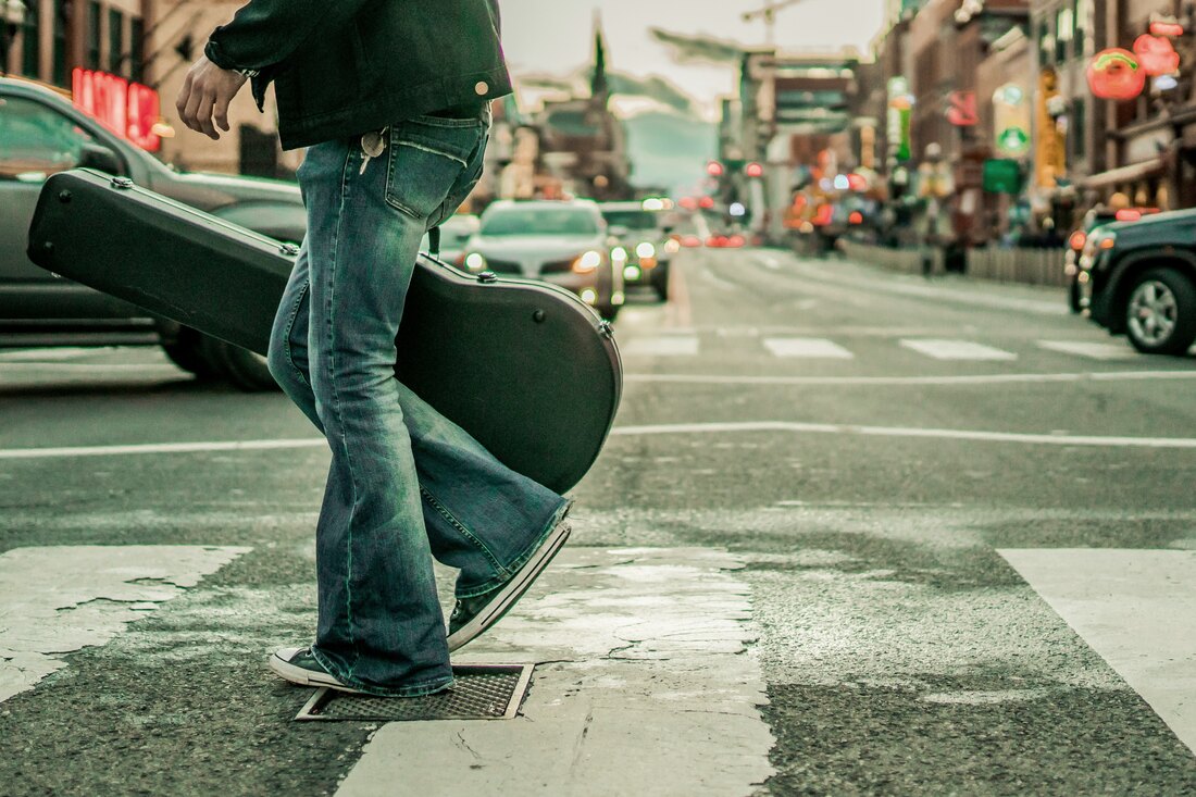 A musician in a denim jacket crossing a city street with a guitar case