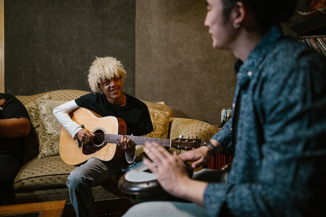 Two musicians on a sofa playing an acoustic guitar and a hand drum in a room lined with acoustic panels