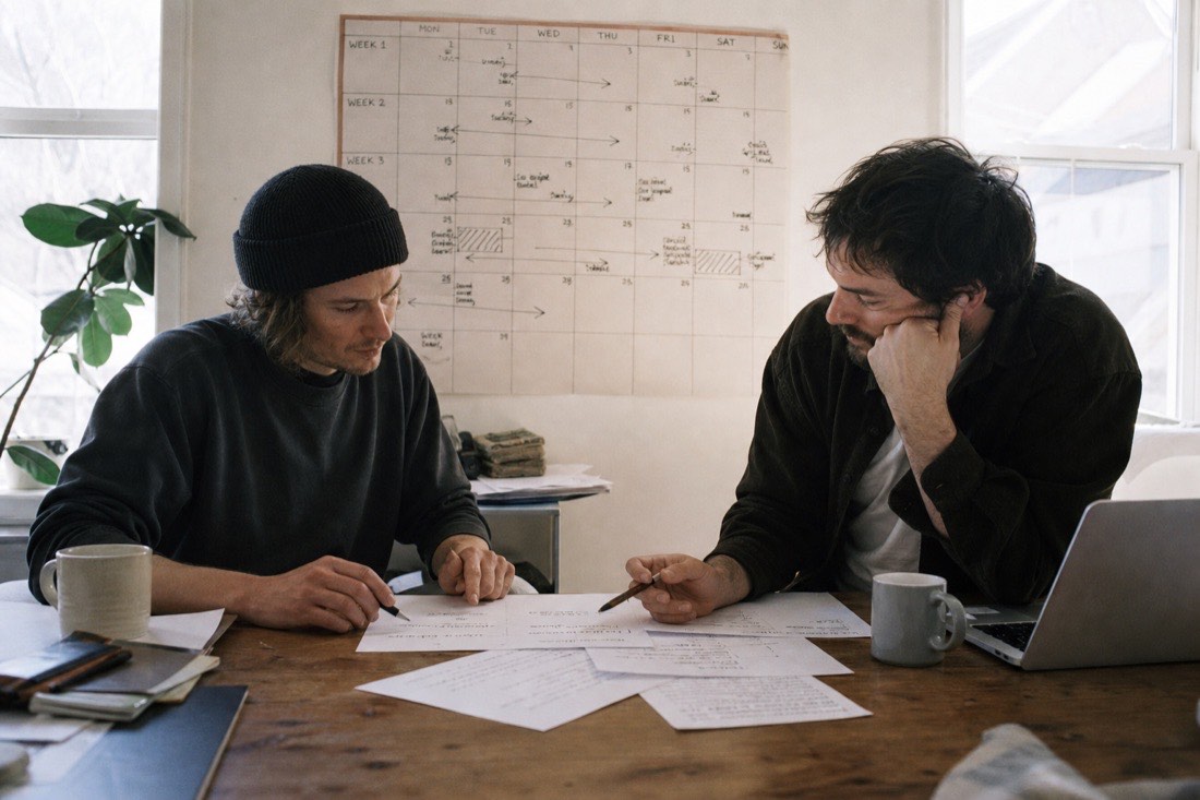 Two musicians at a table working through release paperwork under a hand-drawn three-week wall calendar
