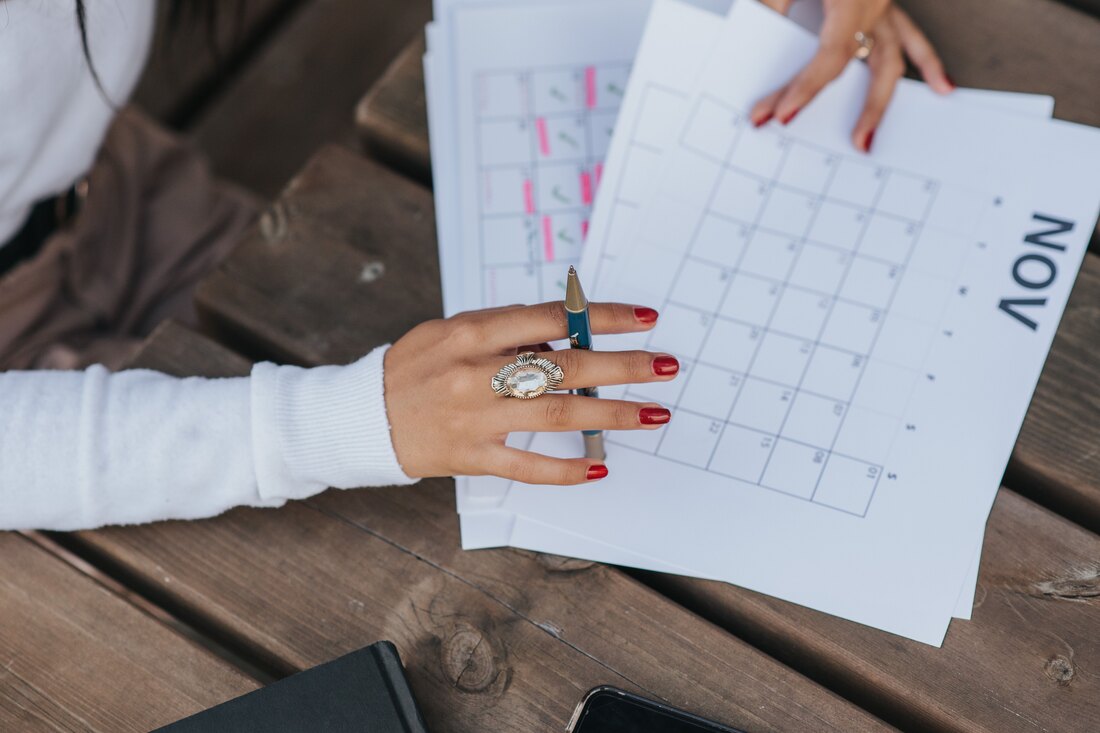 A hand with a pen over a printed month calendar on a wooden table